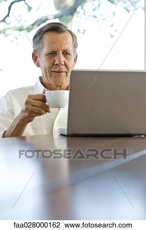 Senior man sitting at table, looking at laptop computer, holding coffee cup View Large Photo Image Stock Image - Senior man sitting at table, looking at laptop computer, holding coffee cup. Fotosearch
