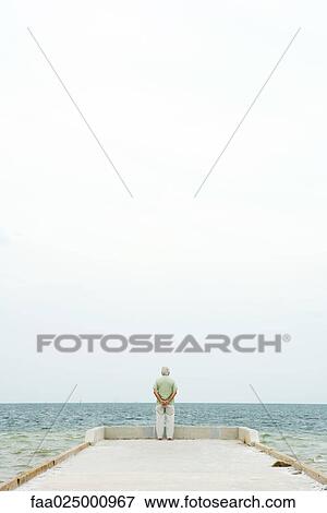 Stock Photo - Senior man standing at end of pier, facing ocean, in distance, rear view. Fotosearch