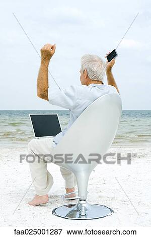 Senior man using laptop and cell phone on beach, raising arms View Large Photo Image Stock Photo - Senior man using laptop and cell phone on beach, raising arms. Fotosearch