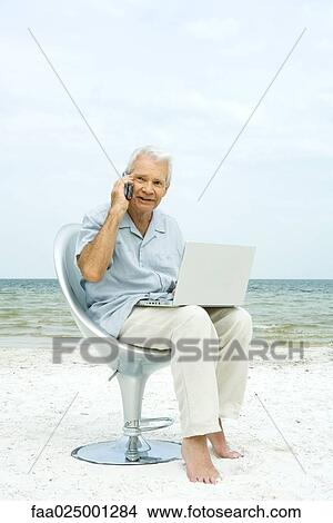 Senior man using laptop on beach, making phone call View Large Photo Image Picture - Senior man using laptop on beach, making phone call. Fotosearch