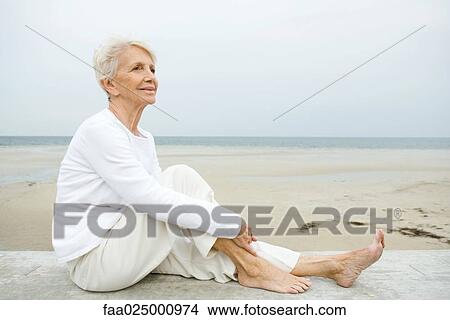 Senior woman sitting on low wall, beach in background, full length View Large Photo Image Picture - Senior woman sitting on low wall, beach in background, full length. Fotosearch