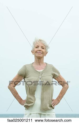 Stock Image - Senior woman standing with hands on hips, smiling at camera, ocean horizon in background, portrait. Fotosearch