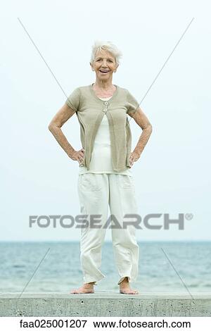 Stock Photo - Senior woman standing with hands on hips, smiling at camera, ocean in background, full length. Fotosearch