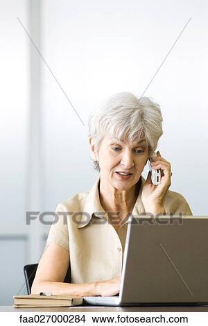 Picture - Senior woman using cell phone and laptop computer, looking down. Fotosearch