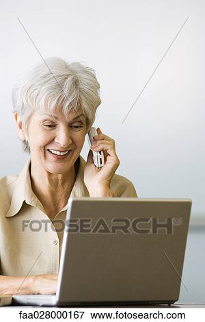 Stock Photo - Senior woman using laptop computer and cell phone, smiling, looking down. Fotosearch