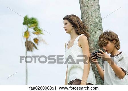 Siblings leaning against tree trunk, boy playing handheld video game, teen girl listening to earphones View Large Photo Image Stock Image - Siblings leaning against tree trunk, boy playing handheld video game, teen girl listening to earphones. Fotosearch