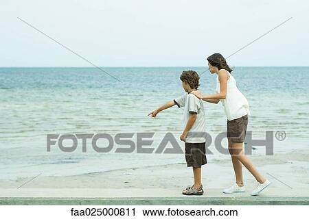 Stock Image - Siblings standing at the beach, boy pointing, both looking together. Fotosearch
