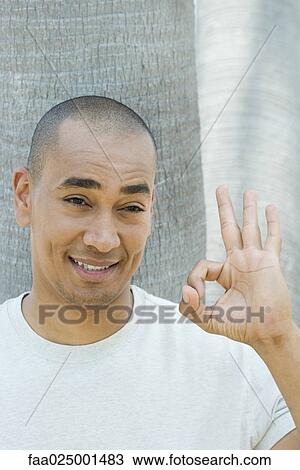 Stock Image - smiling, tree trunk, hand raised, ok sign, raising eyebrows, mid adult men. Fotosearch