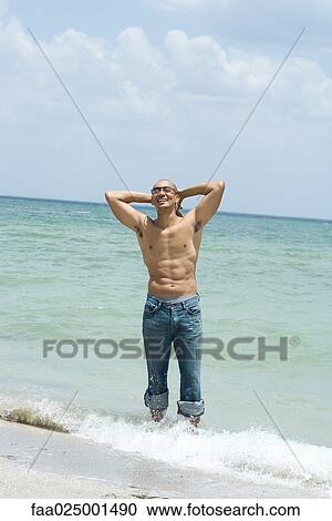Stock Image - standing, barechested, ankle deep in water, hands behind head, water's edge, at the beach. Fotosearch