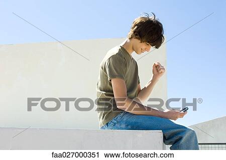 Teenage boy sitting outdoors, looking down at cell phone, making a fist, smiling View Large Photo Image Stock Image - Teenage boy sitting outdoors, looking down at cell phone, making a fist, smiling. Fotosearch