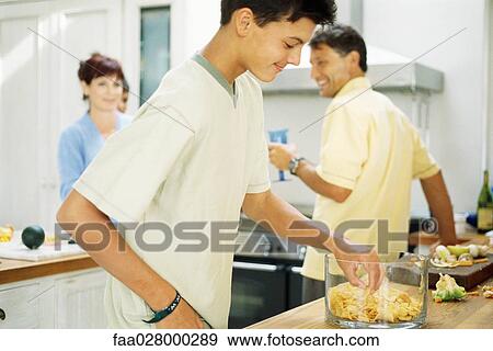 Teenage boy standing in kitchen, eating chips from bowl, family preparing food in the background View Large Photo Image Stock Photo - Teenage boy standing in kitchen, eating chips from bowl, family preparing food in the background. Fotosearch