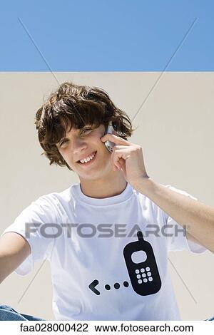Stock Image - Teenage boy wearing tee-shirt printed with cell phone graphic, using cell phone, smiling at camera. Fotosearch