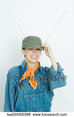 Teenage girl dressed in overalls, putting on hat, smiling at camera View Large Photo Image Stock Photo - Teenage girl dressed in overalls, putting on hat, smiling at camera. Fotosearch