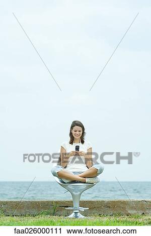 Teenage girl sitting in chair by the sea, looking down at cell phone, smiling View Large Photo Image Stock Image - Teenage girl sitting in chair by the sea, looking down at cell phone, smiling. Fotosearch