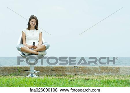 Teenage girl sitting in chair by the sea, holding cell phone, smiling at camera View Large Photo Image Stock Photo - Teenage girl sitting in chair by the sea, holding cell phone, smiling at camera. Fotosearch