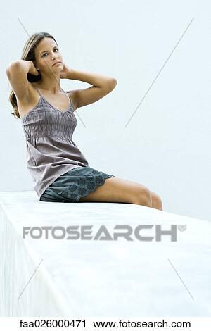 Stock Image - Teenage girl sitting with arms raised, hands in hair, looking at camera. Fotosearch