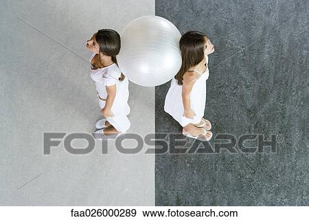 Stock Photo - Teenage twin sisters standing back to back, supporting fitness ball between them, high angle view. Fotosearch
