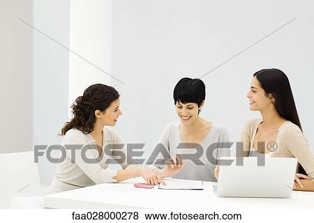 Three businesswomen sitting at table, discussing document, smiling View Large Photo Image Stock Photo - Three businesswomen sitting at table, discussing document, smiling. Fotosearch