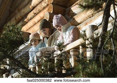 Stock Image - Three preteen or teen girls standing on deck of log cabin, looking away, low angle view. Fotosearch