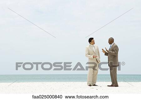 Stock Image - Two businessman standing on beach having conversation. Fotosearch