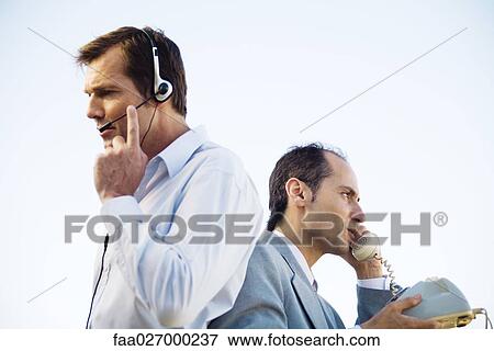 Two businessmen back to back, one using headset, the other using landline phone, side view View Large Photo Image Stock Photo - Two businessmen back to back, one using headset, the other using landline phone, side view. Fotosearch