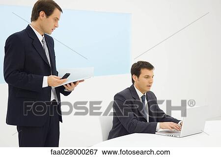 Two businessmen in office looking down at laptop computer together, one holding cell phone View Large Photo Image Stock Photo - Two businessmen in office looking down at laptop computer together, one holding cell phone. Fotosearch
