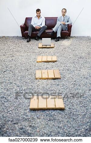 Two businessmen sitting on couch in gravel, looking down at laptop computer, mid-distance View Large Photo Image Stock Photo - Two businessmen sitting on couch in gravel, looking down at laptop computer, mid-distance. Fotosearch