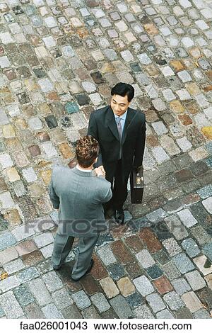 Stock Image - Two businessmen standing face to face, shaking hands, high angle view. Fotosearch