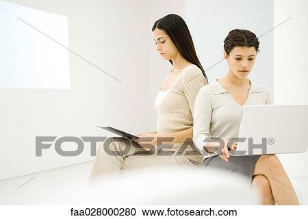 Two businesswomen sitting together, one using laptop computer, the other looking at document View Large Photo Image Stock Image - Two businesswomen sitting together, one using laptop computer, the other looking at document. Fotosearch