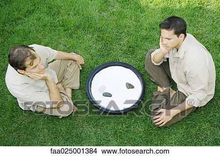 Two men sitting on the ground next to rock garden, both with hands under chins, high angle view View Large Photo Image Picture - Two men sitting on the ground next to rock garden, both with hands under chins, high angle view. Fotosearch