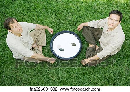Stock Image - Two men sitting on the ground with rock garden between them, looking at camera, high angle view. Fotosearch