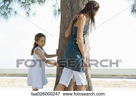Stock Photo - Two sisters standing beside tree trunk, one holding on to the other's shirt with eyes closed. Fotosearch