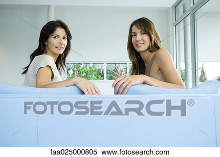 Two women sitting on couch together, both looking over shoulders at camera View Large Photo Image Stock Photography - Two women sitting on couch together, both looking over shoulders at camera. Fotosearch