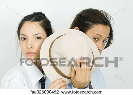 Two young female friends hiding behind hat, looking at camera View Large Photo Image Stock Image - Two young female friends hiding behind hat, looking at camera. Fotosearch