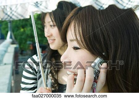 Two young women standing under parasol, one using cell phone, cropped view View Large Photo Image Stock Photography - Two young women standing under parasol, one using cell phone, cropped view. Fotosearch