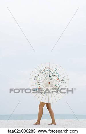 Stock Image - Woman behind parasol walking on beach, full length. Fotosearch