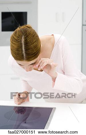Stock Image - Woman bending over stove top, looking at her reflection, touching eye. Fotosearch