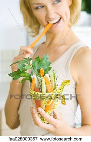 Woman biting into carrot, holding glass full of vegetables tied with measuring tape View Large Photo Image Stock Photo - Woman biting into carrot, holding glass full of vegetables tied with measuring tape. Fotosearch