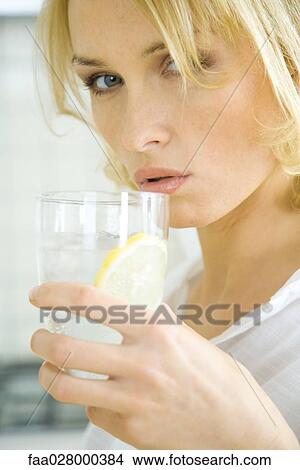 Picture - Woman drinking water with lemon slice, looking at camera, close-up. Fotosearch
