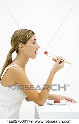 Stock Photo - Woman eating red grape with chopsticks, eyes closed, side view. Fotosearch