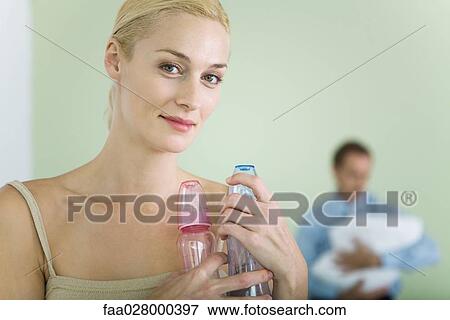 Woman holding baby bottles, smiling at camera, man holding baby in background View Large Photo Image Stock Photo - Woman holding baby bottles, smiling at camera, man holding baby in background. Fotosearch