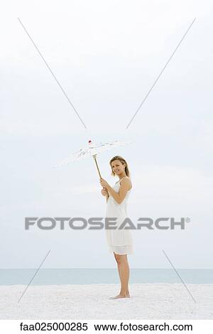 Woman in sundress standing at the beach, holding up parasol, smiling at camera View Large Photo Image Stock Photography - Woman in sundress standing at the beach, holding up parasol, smiling at camera. Fotosearch