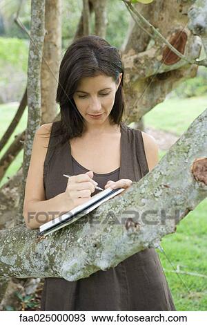 Woman leaning against tree branch, writing in book, looking down View Large Photo Image Stock Image - Woman leaning against tree branch, writing in book, looking down. Fotosearch