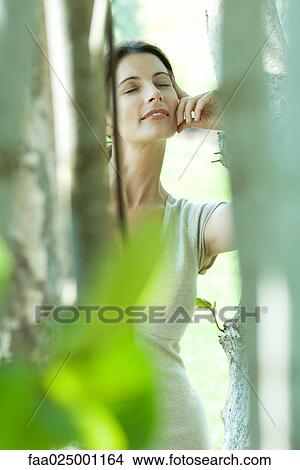 Woman leaning against tree, smiling, eyes closed, view through foliage View Large Photo Image Picture - Woman leaning against tree, smiling, eyes closed, view through foliage. Fotosearch