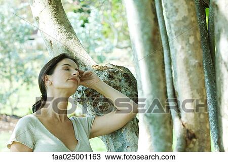 Stock Image - Woman leaning against tree trunk, head back, eyes closed. Fotosearch