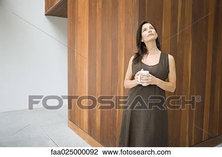 Stock Image - Woman leaning against wall, holding coffee mug, looking up. Fotosearch