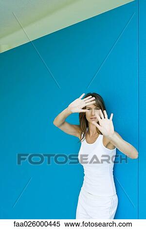 Woman leaning against wall, holding hands up in front of face, looking at camera View Large Photo Image Stock Photography - Woman leaning against wall, holding hands up in front of face, looking at camera. Fotosearch