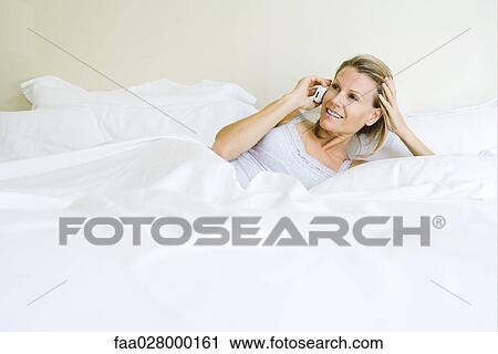 Woman lying in bed, using cell phone, smiling View Large Photo Image Stock Image - Woman lying in bed, using cell phone, smiling. Fotosearch