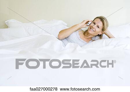 Woman lying in bed, using cell phone, smiling at camera View Large Photo Image Stock Photo - Woman lying in bed, using cell phone, smiling at camera. Fotosearch
