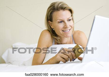 Woman lying in bed, using laptop computer, holding credit card, smiling View Large Photo Image Stock Image - Woman lying in bed, using laptop computer, holding credit card, smiling. Fotosearch